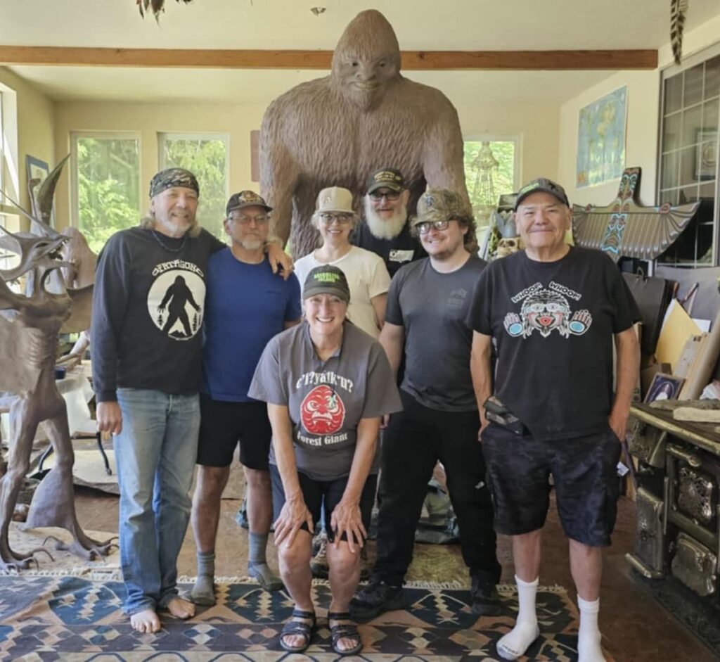 Bigfooting Adventures—Hannah and the Wild Squatch community. Hannah and her husband with a group of Bigfoot enthusiasts, standing in front of a life-sized Bigfoot statue and surrounded by nature-themed decor.