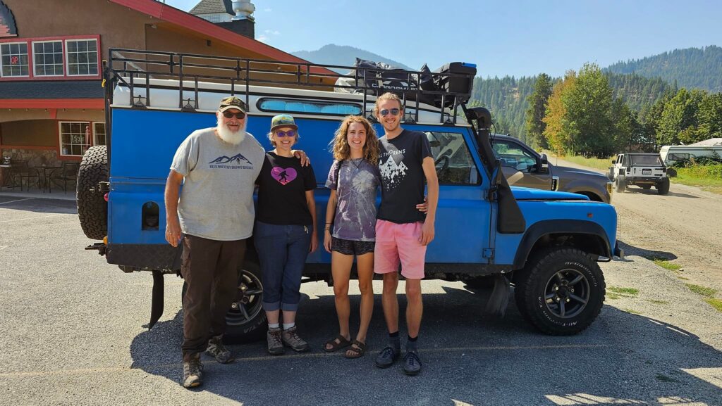 Scot and Hannah and their 2 guests all the way from England, smiling in front of a bright blue off-road vehicle, parked with mountains and trees in the background, celebrating global Squatch America connections.