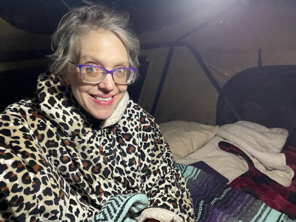 Moonlit tent A woman smiling inside a tent, wrapped in a leopard-print jacket, with moonlight streaming through the tent fly.
