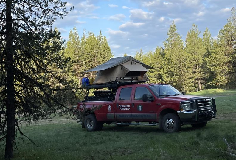 Deep in the woods: A place to reset and recharge off the grid. A red Squatch America truck with a rooftop tent parked deep in the woods, surrounded by tall pine trees and open grassy space under a cloudy sky.