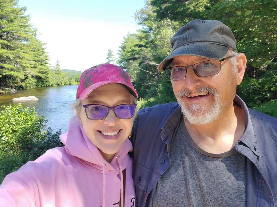 Laughter is the Best Medicine A cheerful couple laughing together by a serene river, surrounded by lush green trees. The woman wears a pink hoodie and matching pink cap, and the man wears a gray shirt and dark cap. Their genuine smiles convey joy and connection.