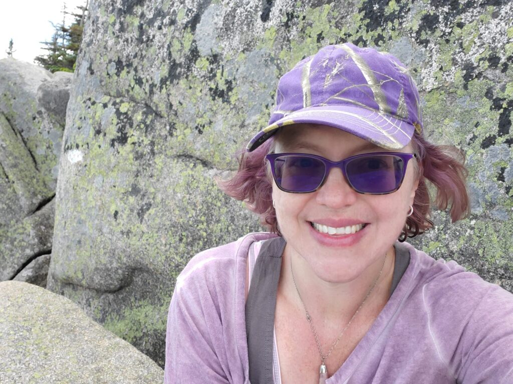 Perched above the treeline on Mt. Katahdin: Rediscovering wonder, joy, and my strength. Hannah, a woman, smiling in a purple hat and glasses, sitting above the treeline on Mt. Katahdin, leaning against the rock wall of the mountain.